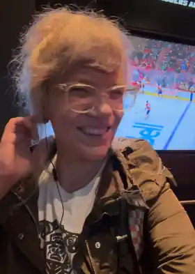 Suzy smiling in translucent glasses, with a hockey game on a screen behind her.